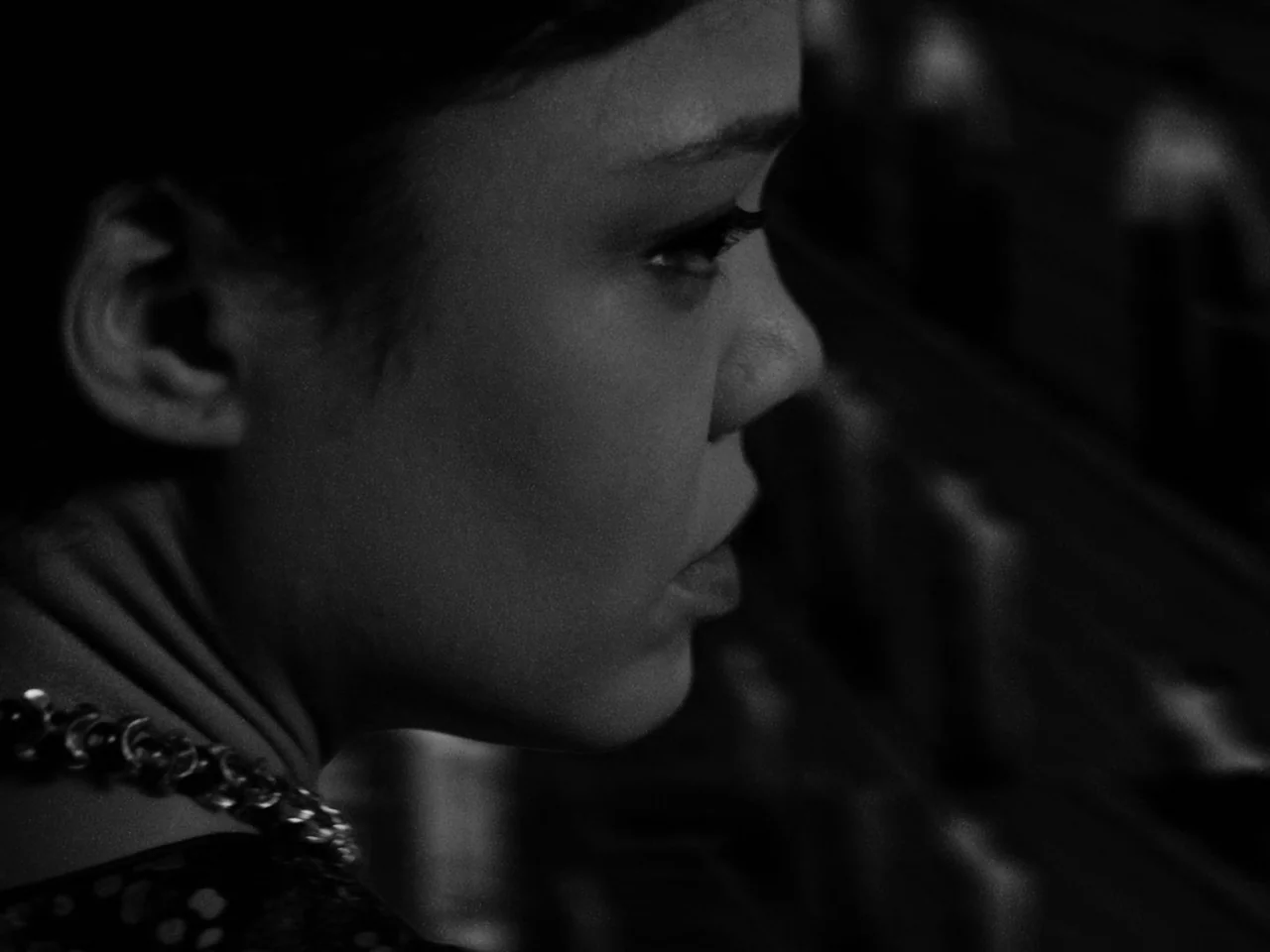 Passing (2021) movie still: closeup — The side profile of a woman is centered against a dark, out-of-focus background. She remains still with a…