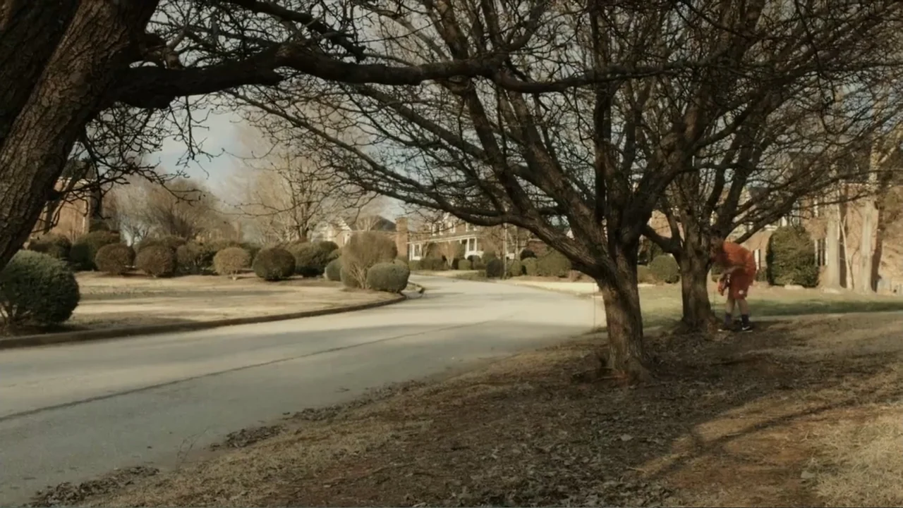 Death of a Child (2017) movie still: wide — A suburban street curves through a neighborhood with leafless trees and manicured lawns. A person in an…