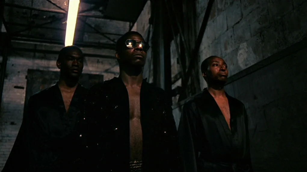 The Warriors (1979) movie still: medium — An industrial space with exposed concrete walls and metal rafters looms in the darkness. Three men in open…