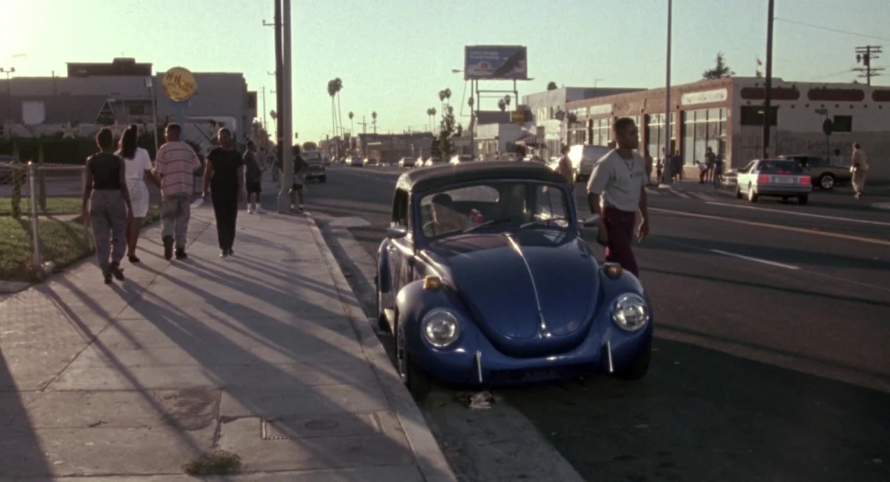 Boyz In The Hood (1991) movie still: wide — A suburban urban street in the early 1990s features a blue Volkswagen beetle parked. A group of people…