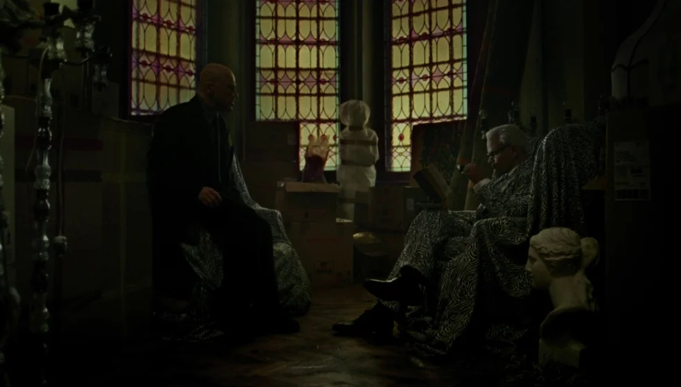 The Zero Theorem (2013) movie still: wide — A dimly lit, cluttered room features stained glass windows and piles of cardboard boxes. Two men sit in…