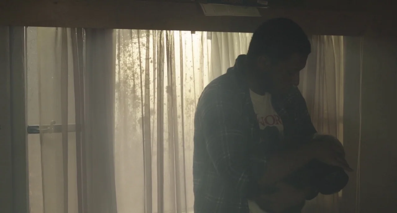 The Alchemist Cookbook (2016) movie still: medium — An interior space features a window covered by thin, textured curtains. A young man stands in…