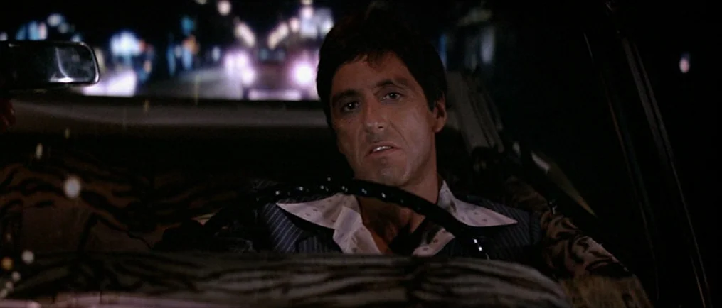 Scarface (1983) movie still: medium — The interior of a car at night with a blurred cityscape visible through the windshield. A man wearing a…
