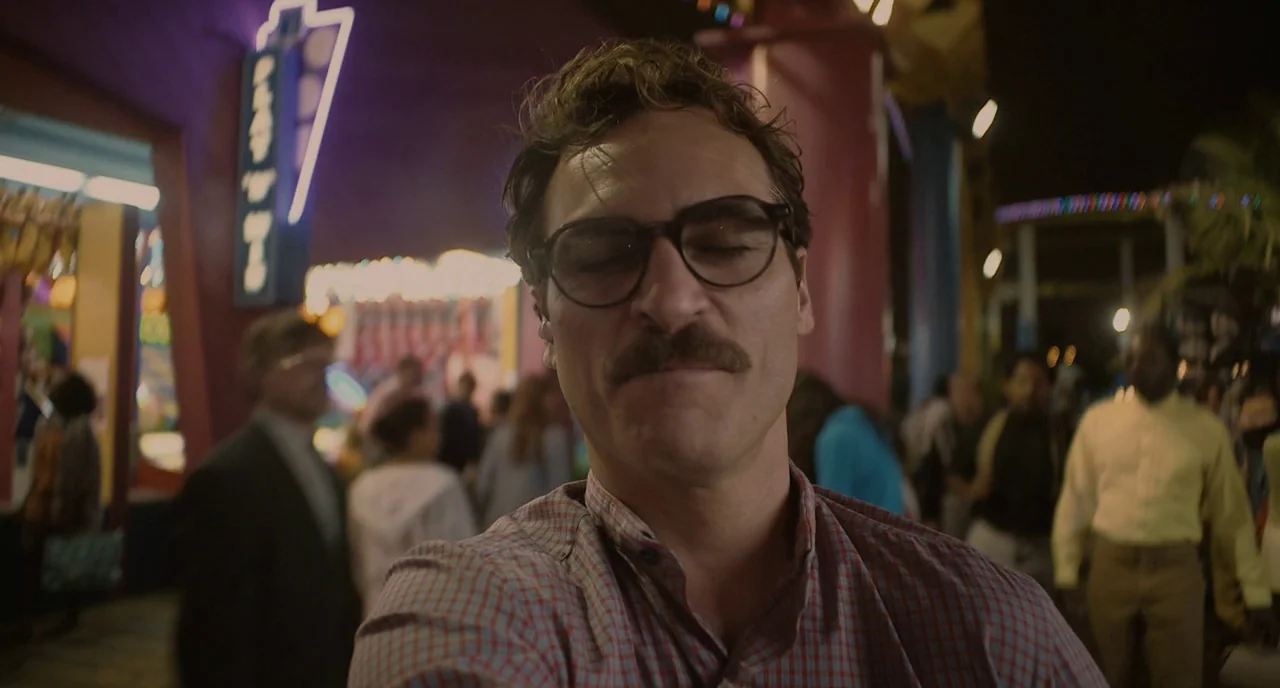 Her (2013) movie still: closeup — A nighttime amusement park environment filled with blurred neon lights and distant moving people. A man in a plaid…
