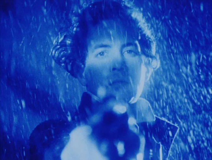 Careful (1992) movie still: closeup — A man stands outdoors at night during a heavy downpour of rain. He holds a firearm directed towards the lens…