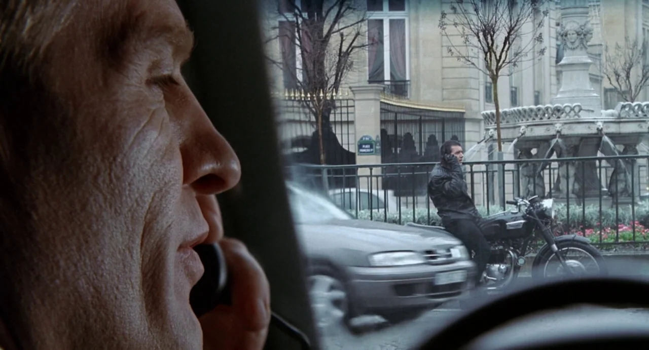 Femme Fatale (2002) movie still: medium — A man in the driver seat speaks on a phone while observing the street. In the background a motorcyclist…