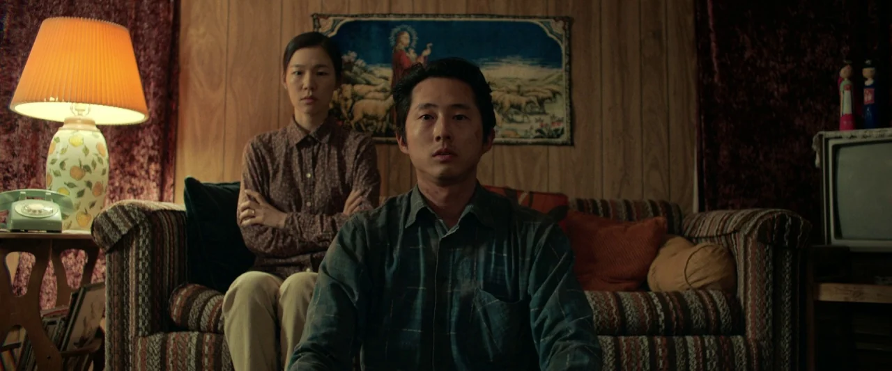 Minari (2020) movie still: medium — A 1980s style space with wood paneling and a striped vintage sofa. A man and a woman sit together looking…