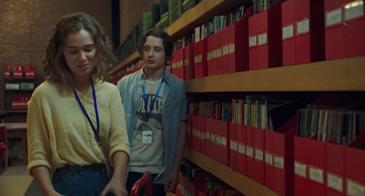 Columbus (2017) movie still: medium — A long aisle of tall shelves filled with red cardboard files in an archive. A young woman and man walk past…