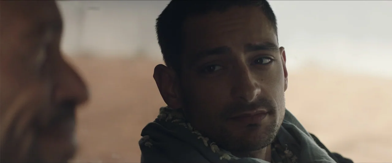 Redemption Day (2021) movie still: closeup — A sun-drenched, sandy outdoor area with blurred background elements. A man wearing a scarf looks…