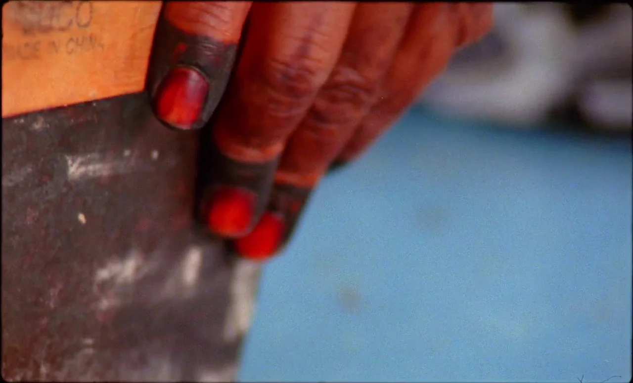 Samsara (2023) movie still: closeup — A close view shows fingers with red-stained cuticles resting against a metal object. The background consists…