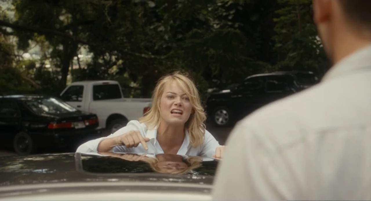 Aloha (2015) movie still: medium — A sunny outdoor parking lot with multiple vehicles parked under mature leafy trees. A woman leans over the roof…