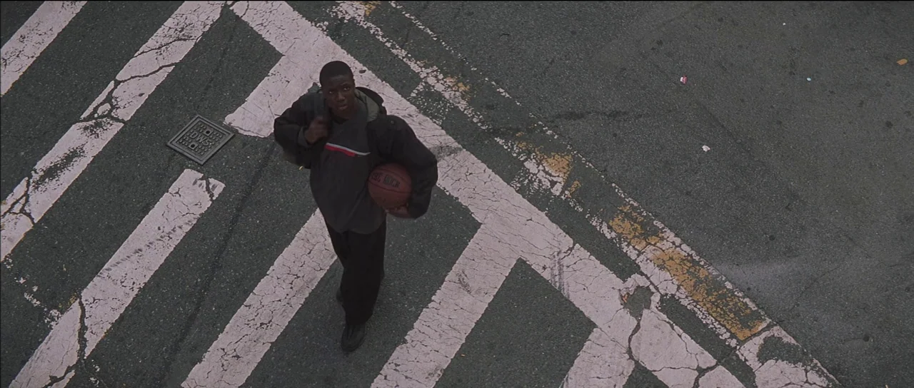 Finding Forrester (2000) movie still: wide — An asphalt street features worn white painted lines of a pedestrian crosswalk. A young man walks across…