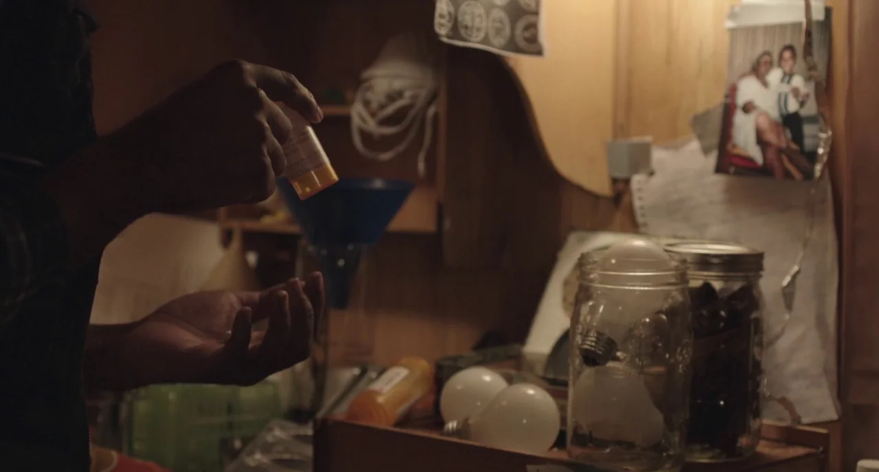 The Alchemist Cookbook (2016) movie still: closeup — A cluttered shelf in a dimly lit domestic space features jars and light bulbs. A hand holds a…