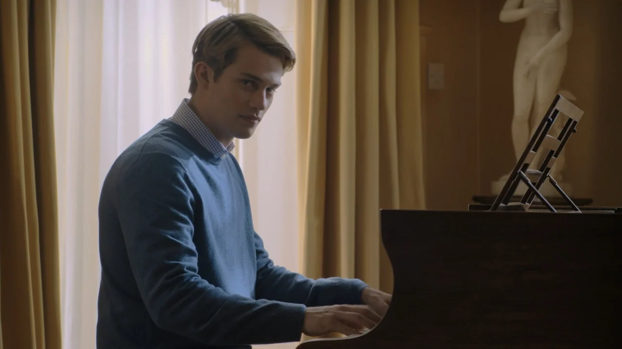 Red, White & Royal Blue (2023) movie still: medium — A well-lit room features a piano positioned near floor-to-ceiling draped curtains. A young man…