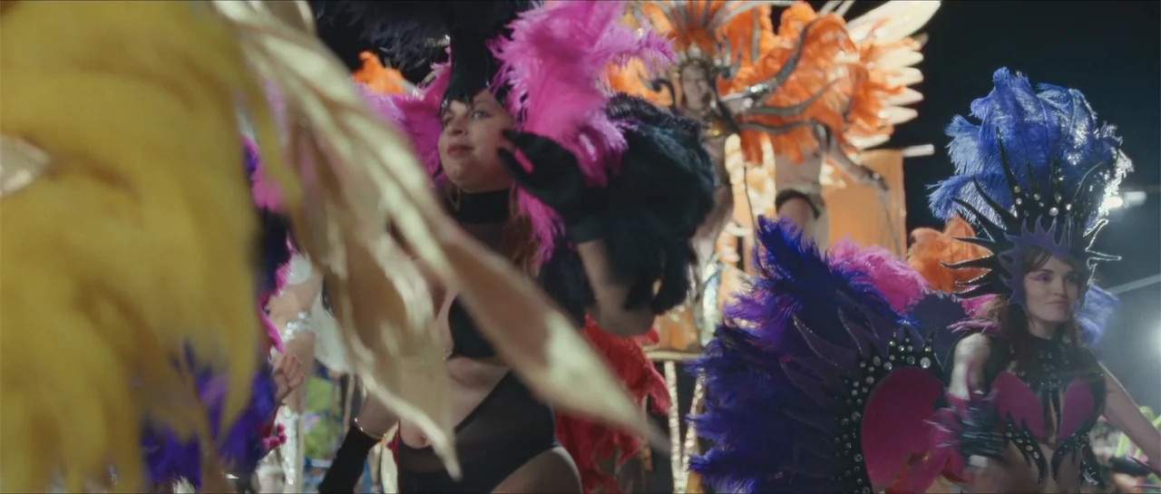 Nahir (2024) movie still: medium — A group of carnival performers wearing large feather headdresses and elaborate costumes walk forward. They move…