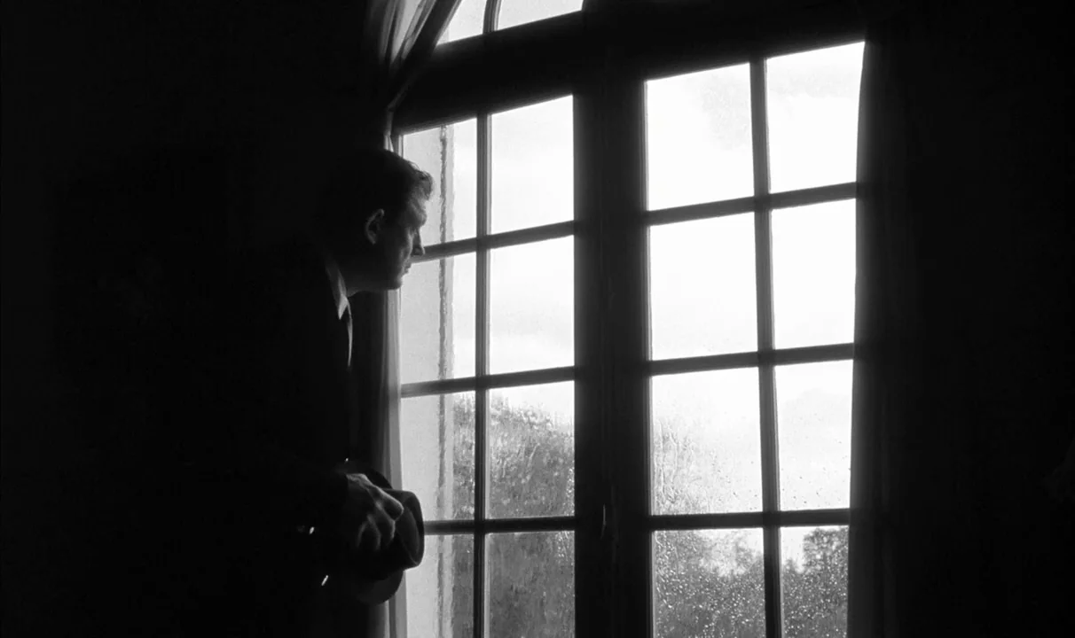 Le Doulos (1962) movie still: medium — A man stands near a large paneled window with rain streaks on the glass. He holds a hat in his hand while…