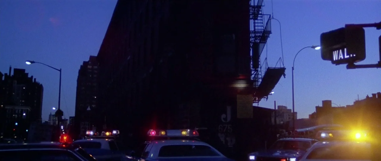 The New York Ripper (1982) movie still: wide — A dark city street is lined with multi-story brick buildings during the blue hour. Several police…