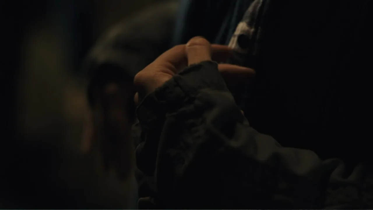 Paterno (2018) movie still: closeup — A dark indoor space with minimal light reveals a close-up of a hand. A person wearing a dark jacket over a…