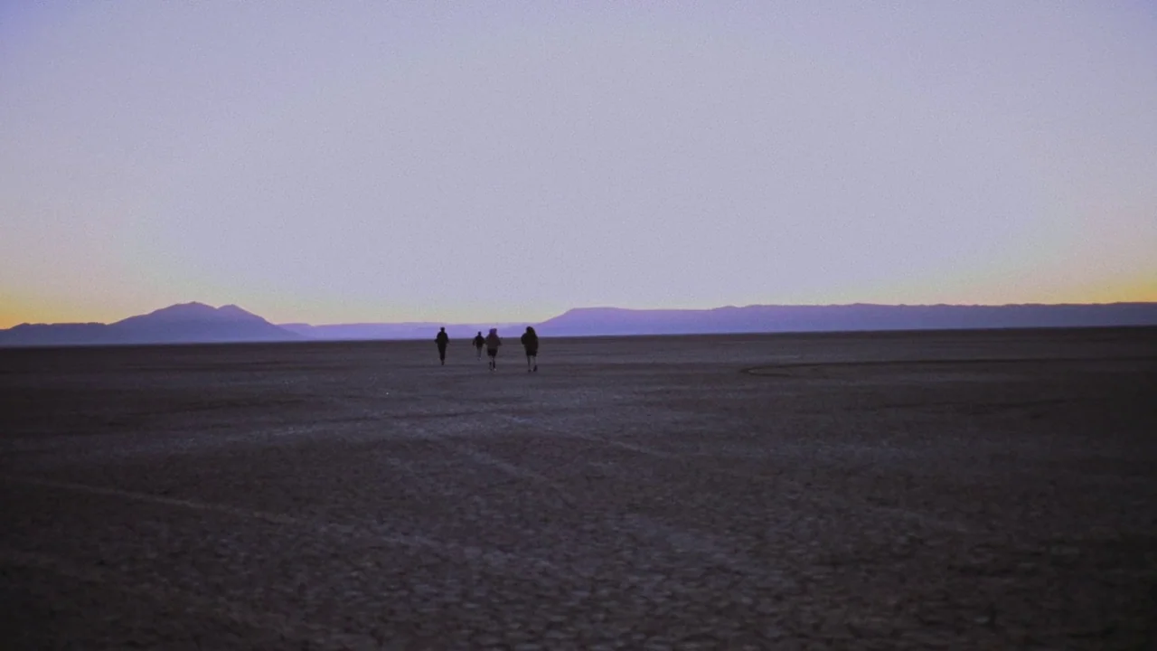 Gasoline Rainbow (2023) movie still: wide — A vast, flat, and arid desert floor extends to a distant mountain range. Three small figures walk away…