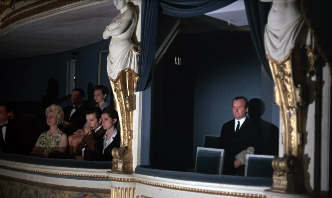 The Bride Wore Black (1968) movie still: wide — A formal theater box features ornate gold carved statues and deep blue velvet upholstery. A man in a…