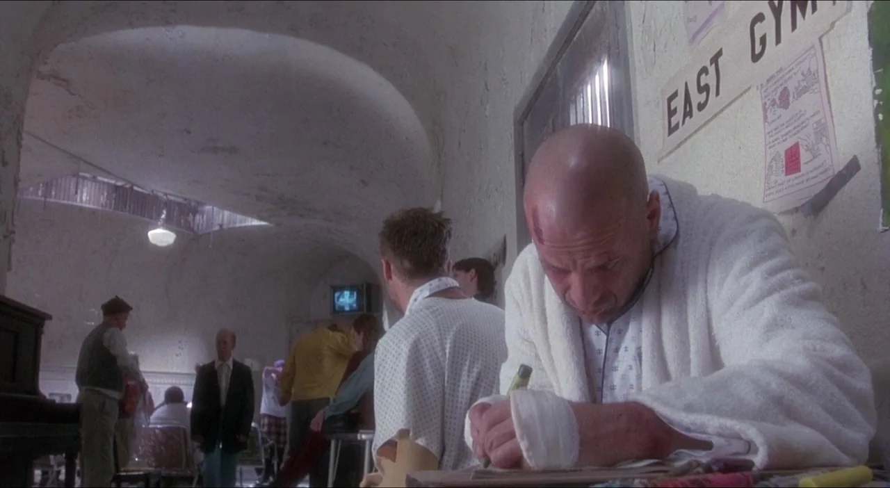 Twelve Monkeys (1995) movie still: medium — A sterile, vaulted hospital common area features textured walls and posters on the pillars. A bald man…