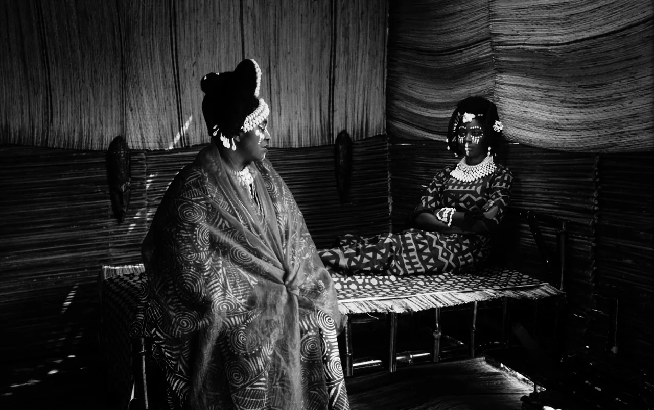 Mami Wata (2023) movie still: fullbody — A dark, textured interior space is constructed from bundled reeds and straw. Two women with white facial…