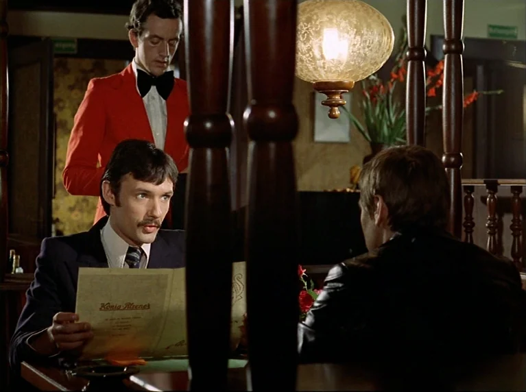 Fox and His Friends (1975) movie still: medium — A dimly lit establishment features dark wood partitions and a low-hanging glass pendant light. A…