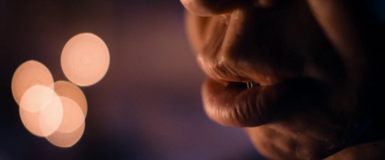 Baskin (2015) movie still: closeup — A dark environment with blurred bokeh light circles in the background. A human mouth is partially lit in side…
