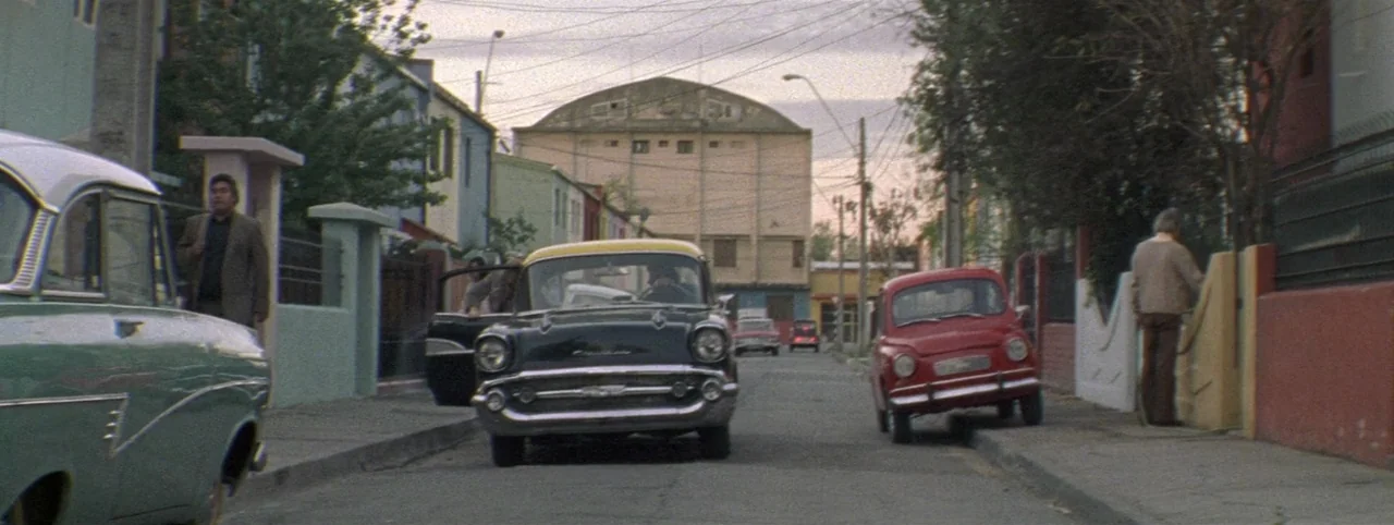 Post Mortem (2010) movie still: wide — A narrow residential street features multi-colored houses and multiple classic vintage automobiles. Men stand…