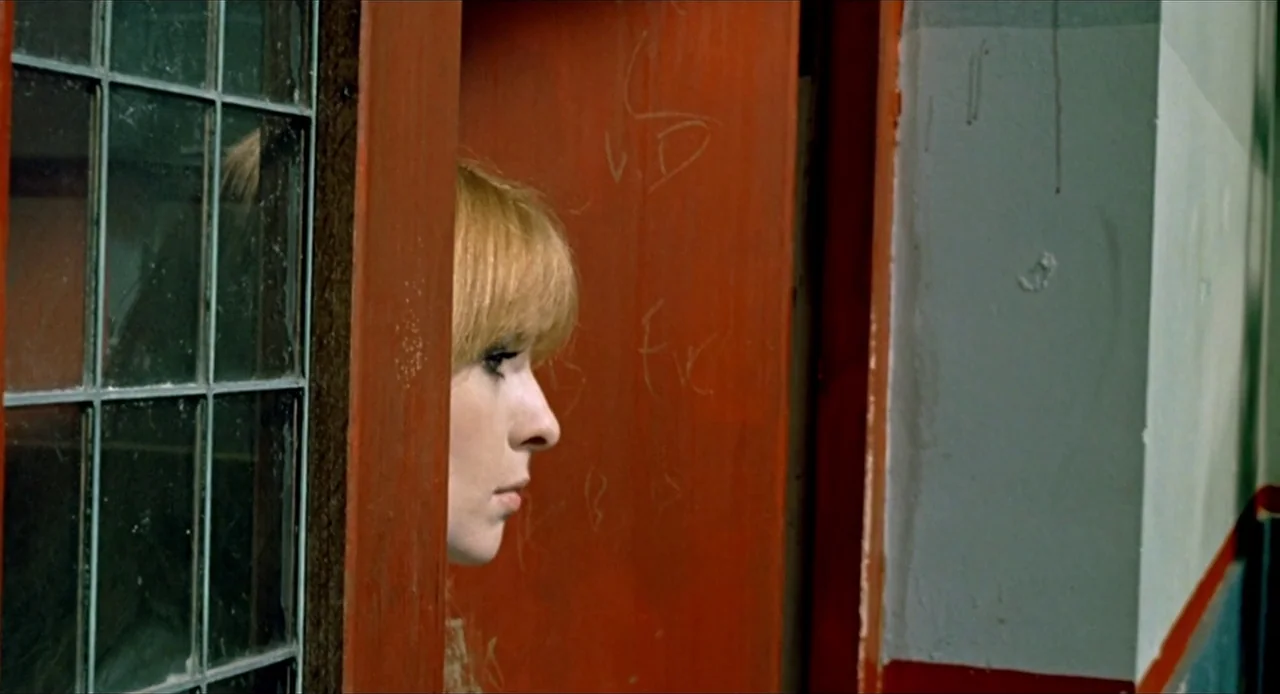 Deep End (1970) movie still: closeup — A stairwell landing features a bright red door and a window with square panes. A woman stands partially…