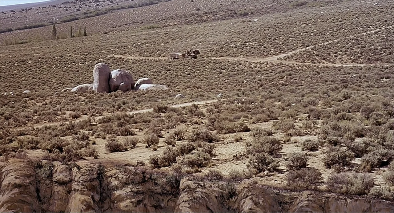 Tremors (1990) movie still: wide — A vast, arid desert landscape features low-lying scrub brush and a few large boulders. A distant dirt trail…