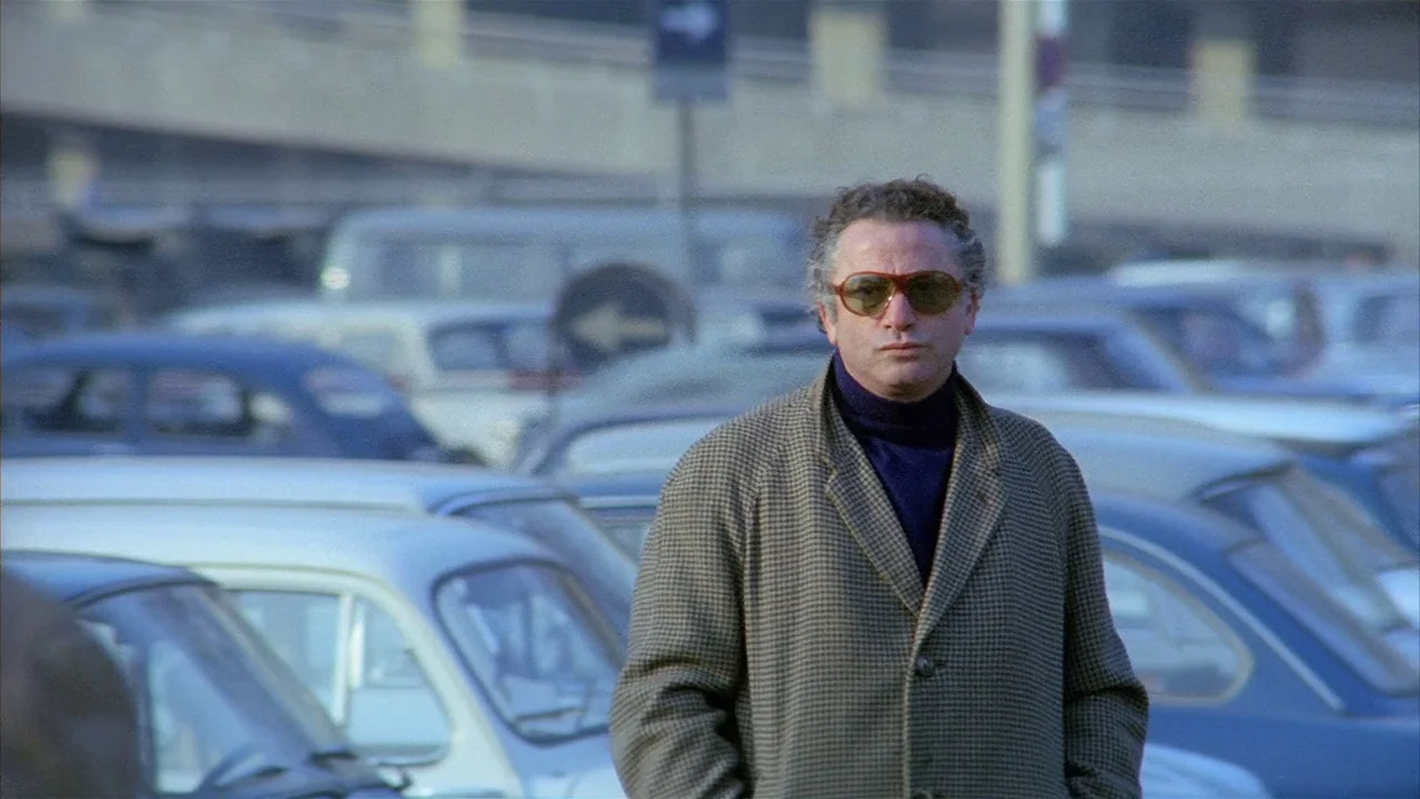 Death Occurred Last Night (1970) movie still: medium — A crowded outdoor parking lot features rows of vintage vehicles in the background. A…