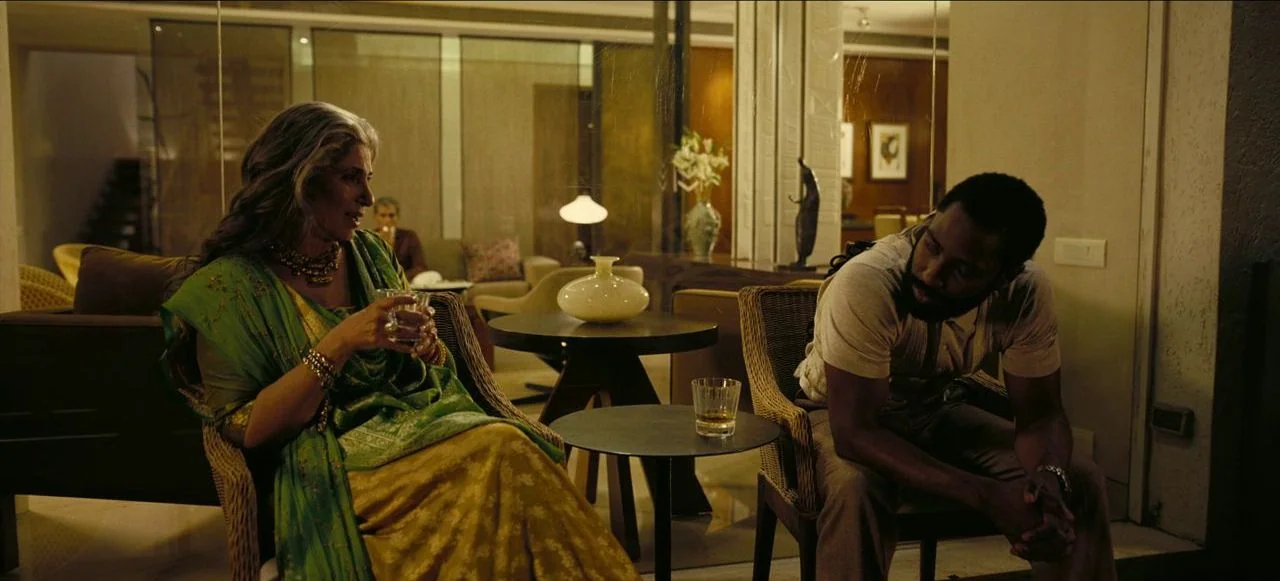 Tenet (2020) movie still: medium — A well-lit contemporary interior space with glass walls and minimalist furniture. A man and a woman sit in chairs…