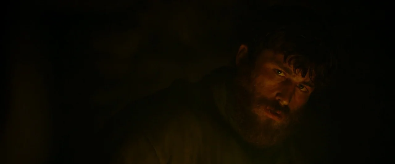 Black ’47 (2018) movie still: closeup — A dark, cavernous space is illuminated only by a low, warm light source. A bearded man looks toward the side…