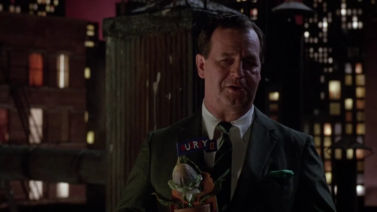 Little Shop of Horrors (1986) movie still: medium — A man stands on a rooftop at night against a blurred urban background. He holds a small plant…