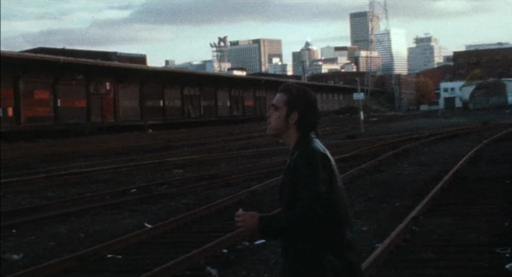 Drugstore Cowboy (1989) movie still: medium — An abandoned industrial railyard sits beneath a distant, sparse urban skyline at twilight. A man…