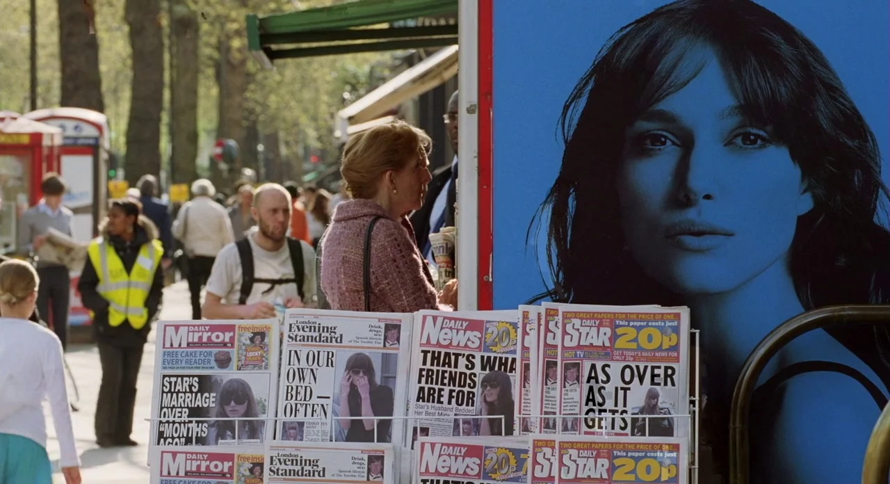London Boulevard (2010) movie still: medium — A bustling outdoor newsstand is positioned on a sidewalk in a busy urban area. A woman in a pink…