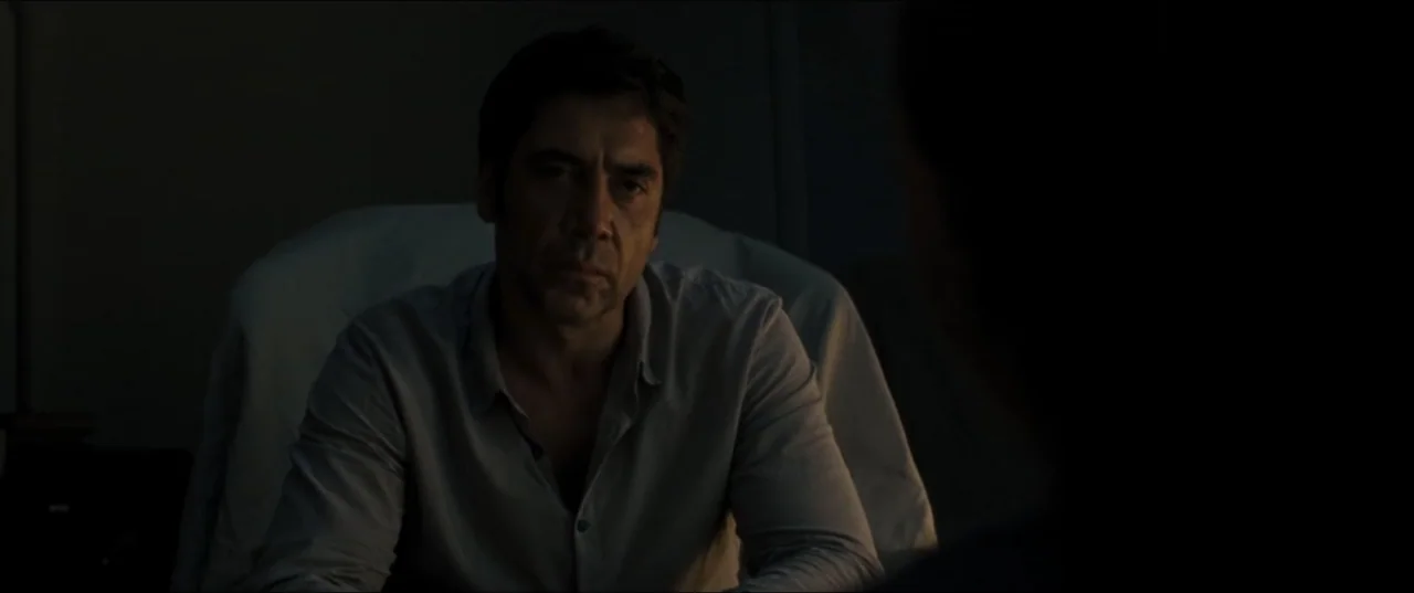 mother! (2017) movie still: medium — A dark, dimly lit office interior contains a man seated at a desk. The man in a grey button-up shirt stares…