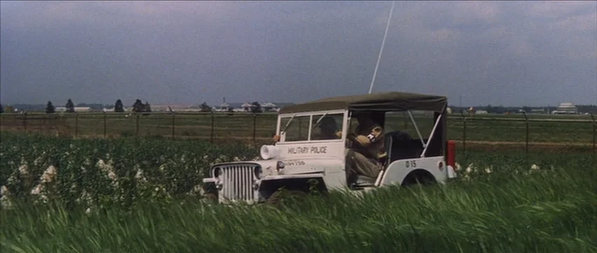 Gate of Flesh (1964) movie still: wide — A white military police jeep drives through a grassy field near a fenced boundary. The vehicle is marked…
