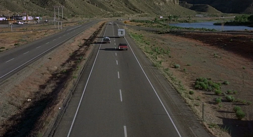 Vanishing Point (1971) movie still: wide — A wide, multi-lane highway stretches through a barren, arid landscape with rolling hills. Three vehicles…