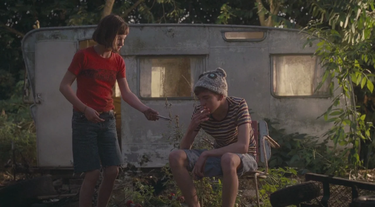Broken (2012) movie still: medium — A weathered vintage trailer sits in an overgrown area with thick foliage and debris. A girl standing offers a…
