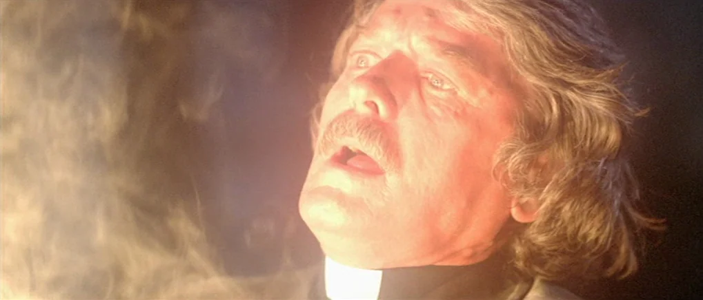 The Fog (1980) movie still: A priest is caught in a blinding, ethereal golden light that cuts through a dark, smoky void. His expression of profound…
