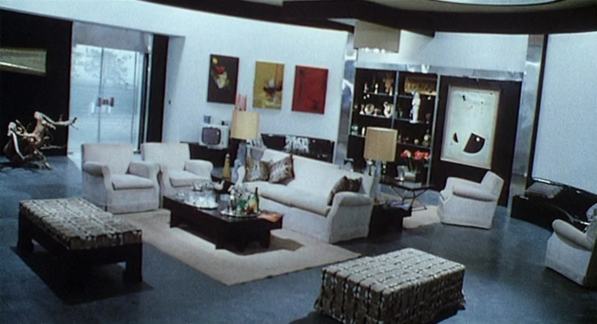 5 Dolls for an August Moon (1970) movie still: wide — A 1960s style open-plan living area features white walls, dark flooring, and modernist…