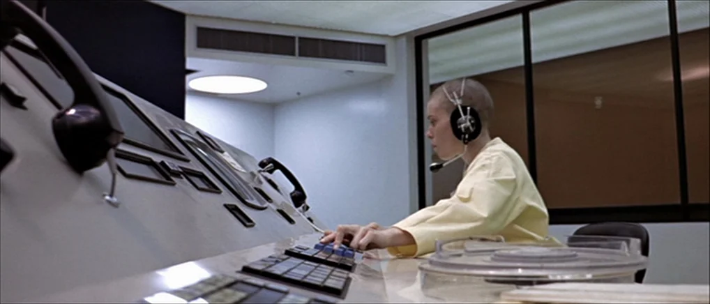 THX 1138 (1971) movie still: medium — A minimalist control room features a large, slanted console with vintage communication equipment. A woman sits…