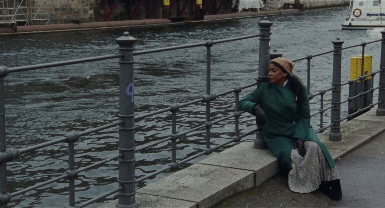 Origin (2023) movie still: fullbody — A stone embankment borders a dark river with a metal security railing. A woman in a green coat sits on the…
