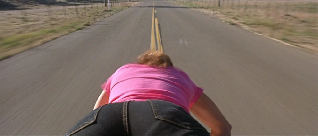 Death Proof movie still: medium — A long stretch of paved country road extends toward the horizon under daylight. A person lies flat on the road…