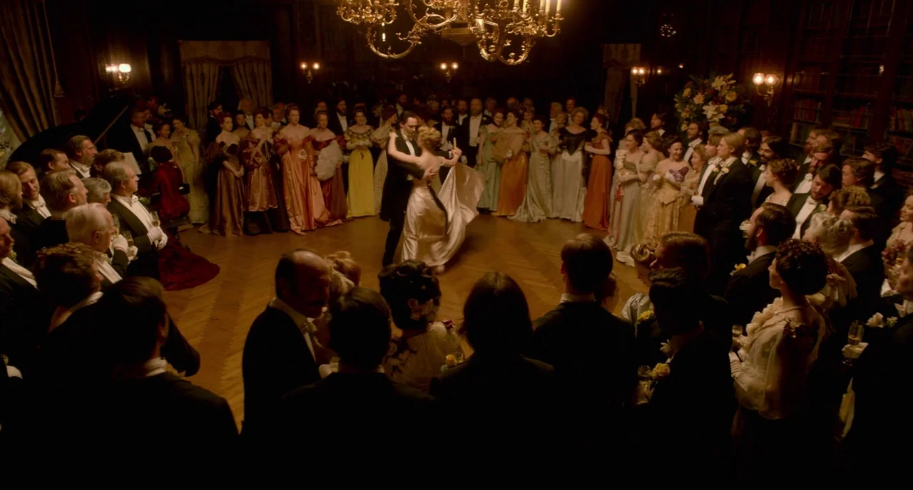 Crimson Peak (2015) movie still: wide — A crowded, dimly lit ballroom features ornate wood-paneled walls and a large gold chandelier. A formally…