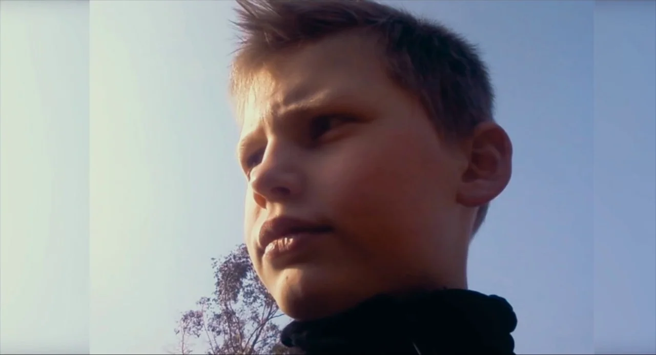 Happy End (2017) movie still: closeup — An outdoor setting features a clear blue sky backdrop. A young boy looks away from the camera in a low-angle…