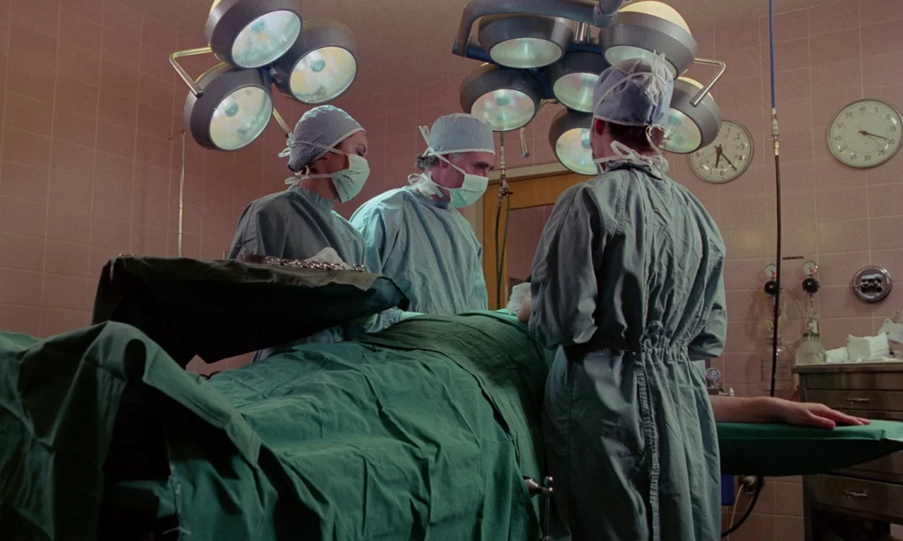 Rabid (1977) movie still: medium — A medical operating room features pink tiled walls and multiple overhead surgical lamps. Three surgeons in blue…