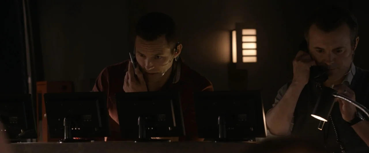 On a Wing and a Prayer (2023) movie still: medium — A dimly lit room features rows of computer terminals and wall-mounted sconces. Two men in work…